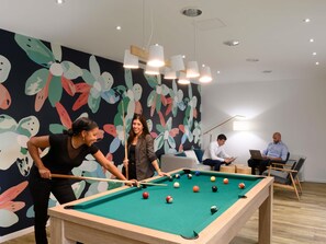Uma mesa de bilhar com bolas, uma pessoa jogando e outra observando. Há luminárias no teto e um mural colorido na parede.