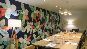 Een vergaderzaal met een lange tafel, stoelen, een bloemistisch wandtapijt en plafondlampen.