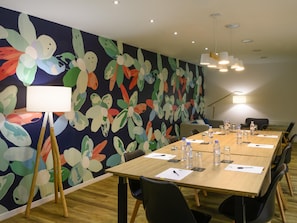 Une salle de conférence avec une longue table, des chaises, une fresque murale à motifs floraux et des luminaires au plafond.