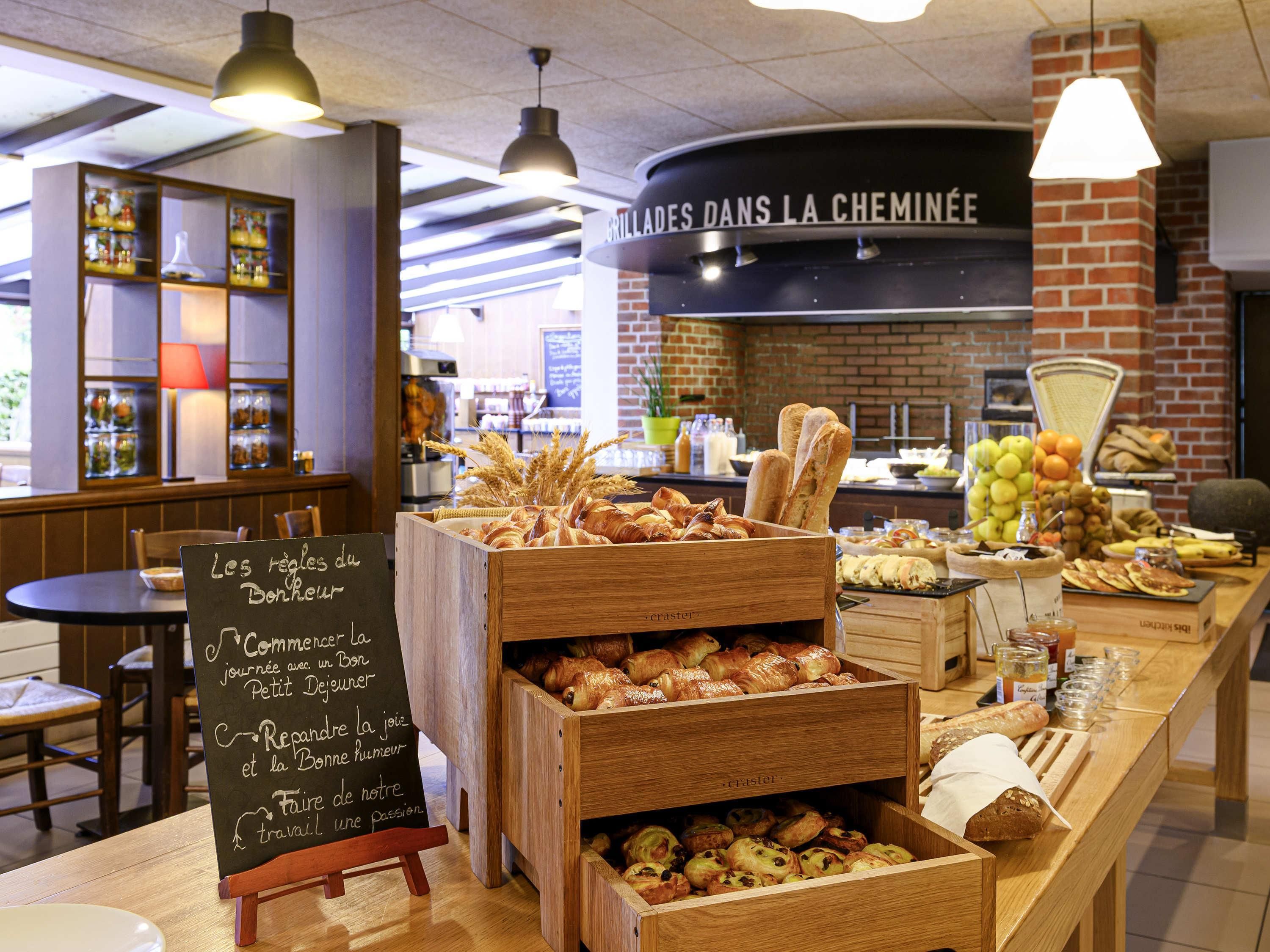 Un comptoir de boulangerie avec du pain et des pâtisseries, un menu écrit à la craie et une pancarte indiquant : « GRILLADES AU FEU DE CHEMINEE ».