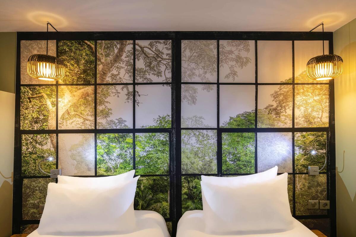 A room with two beds, a large window with a view of trees, and two wall-mounted lamps.
