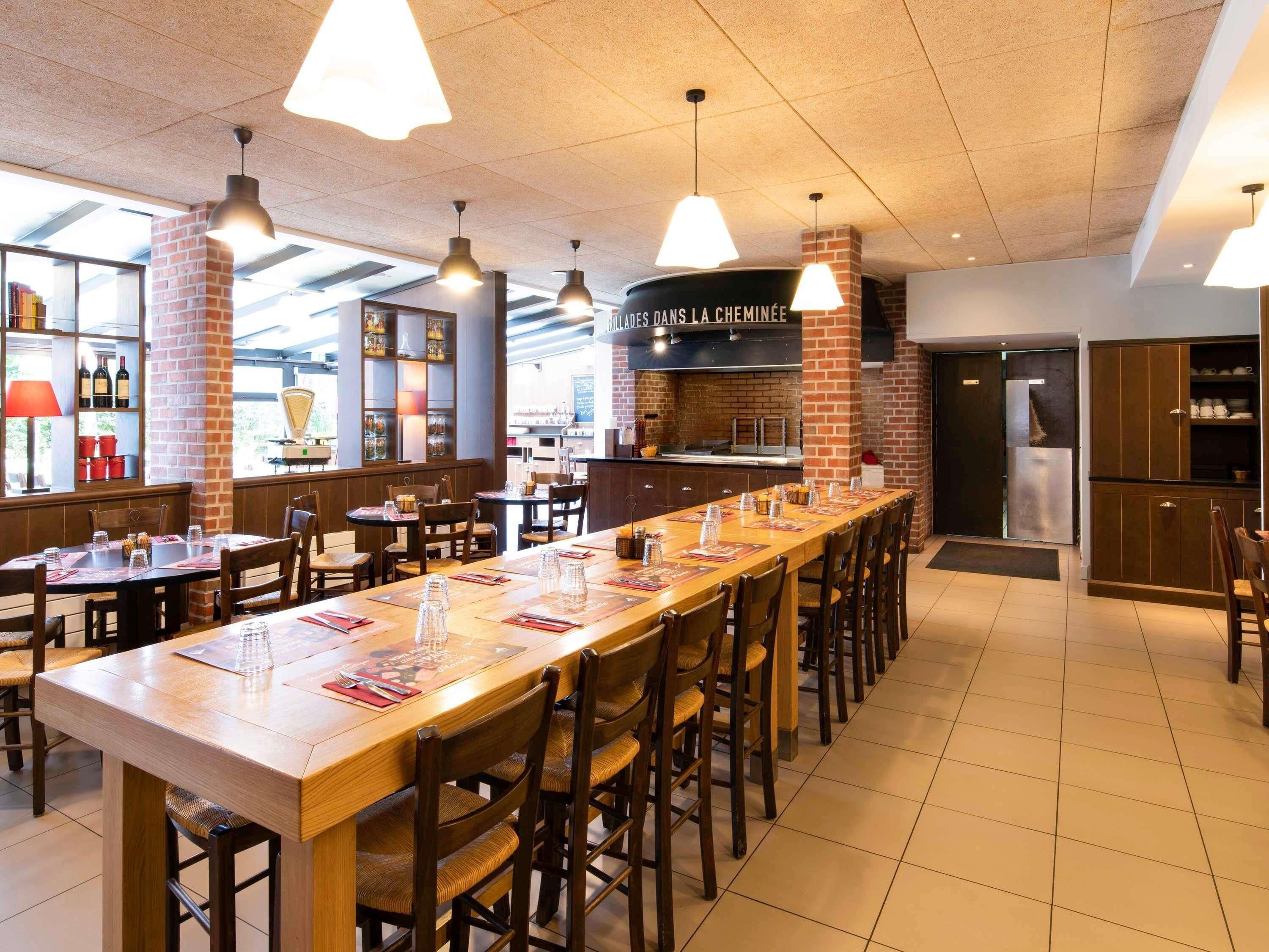 A restaurant with a long wooden counter, multiple tables, and chairs. There are hanging pendant lights, a brick wall, and a kitchen area with a grill.