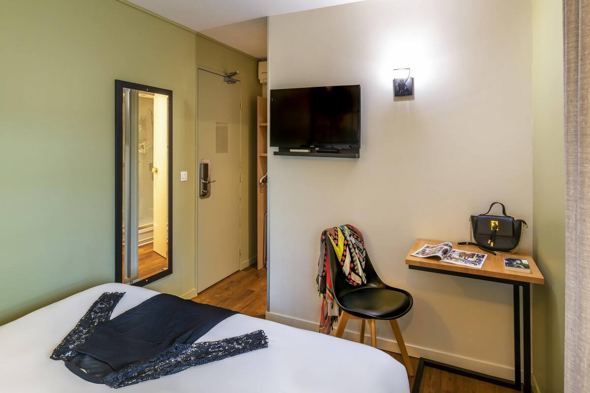 A hotel room with a bed, a desk with a chair, a TV, and a door leading to a bathroom.