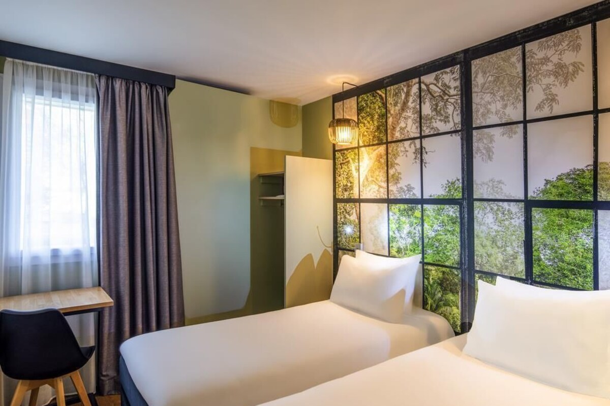A hotel room with two beds, a desk, a chair, and a large window with a view of trees.