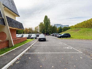 Un estacionamiento con varios coches, un edificio de techo inclinado y una colina cubierta de césped al fondo.