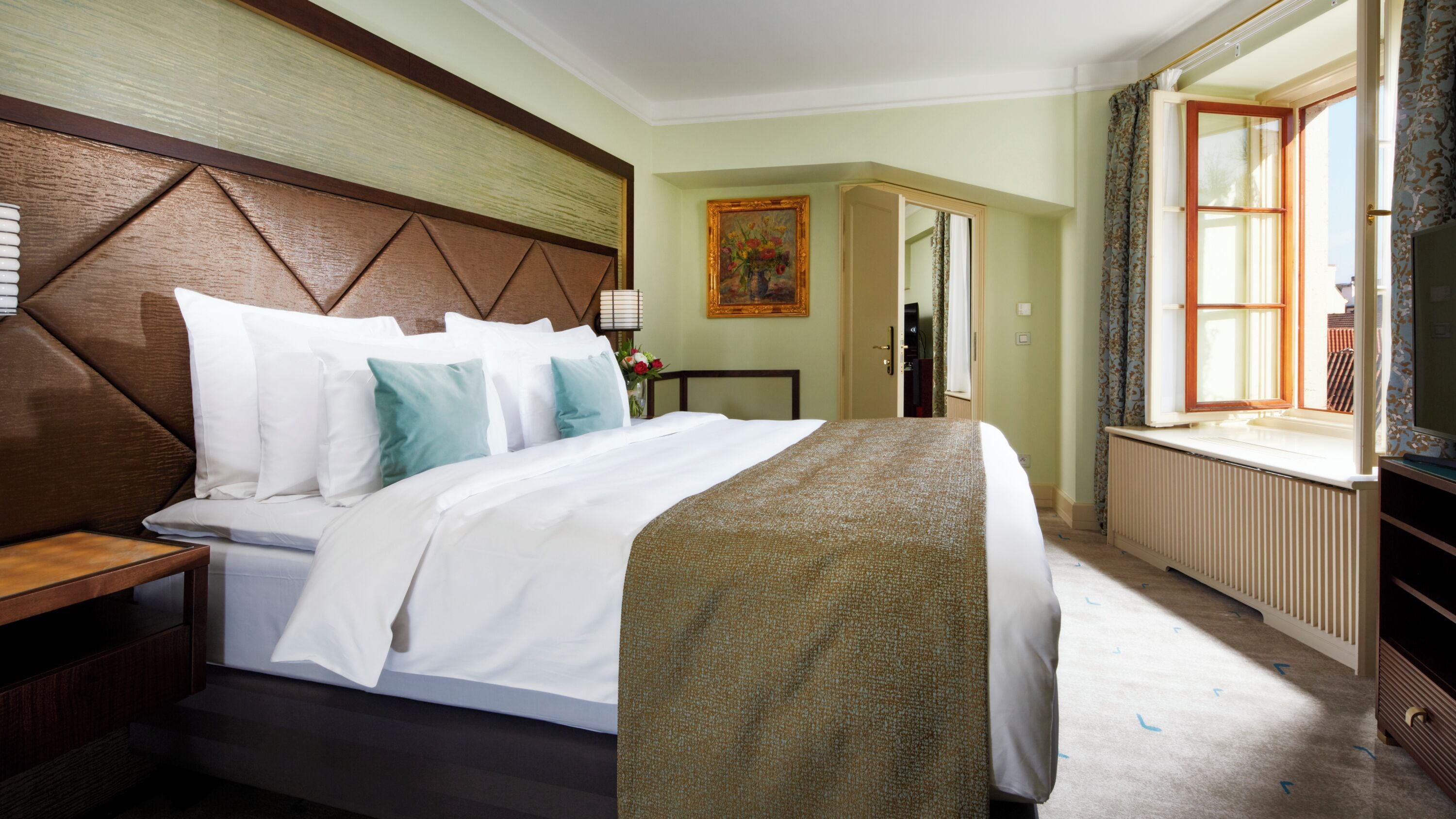 A hotel room with a large bed, a wooden headboard, a bedside table, a nightstand, a window with curtains, a radiator, and a painting on the wall.