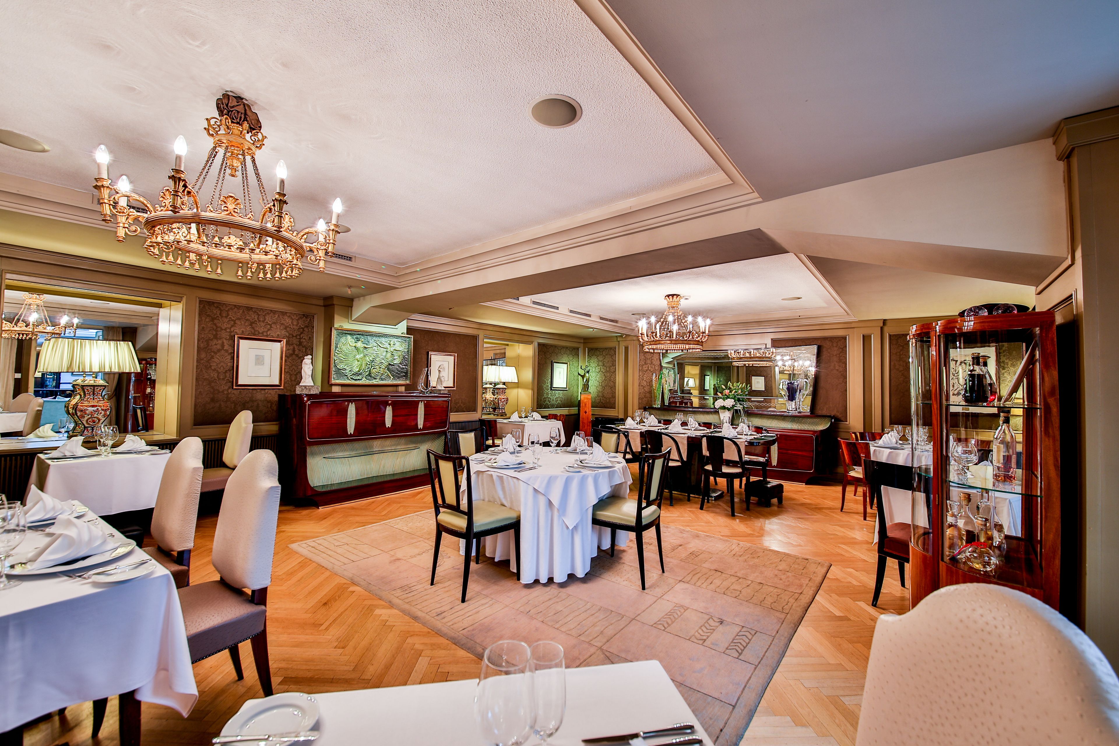 A spacious dining area with multiple tables set for dining, a chandelier, and a wine display cabinet.