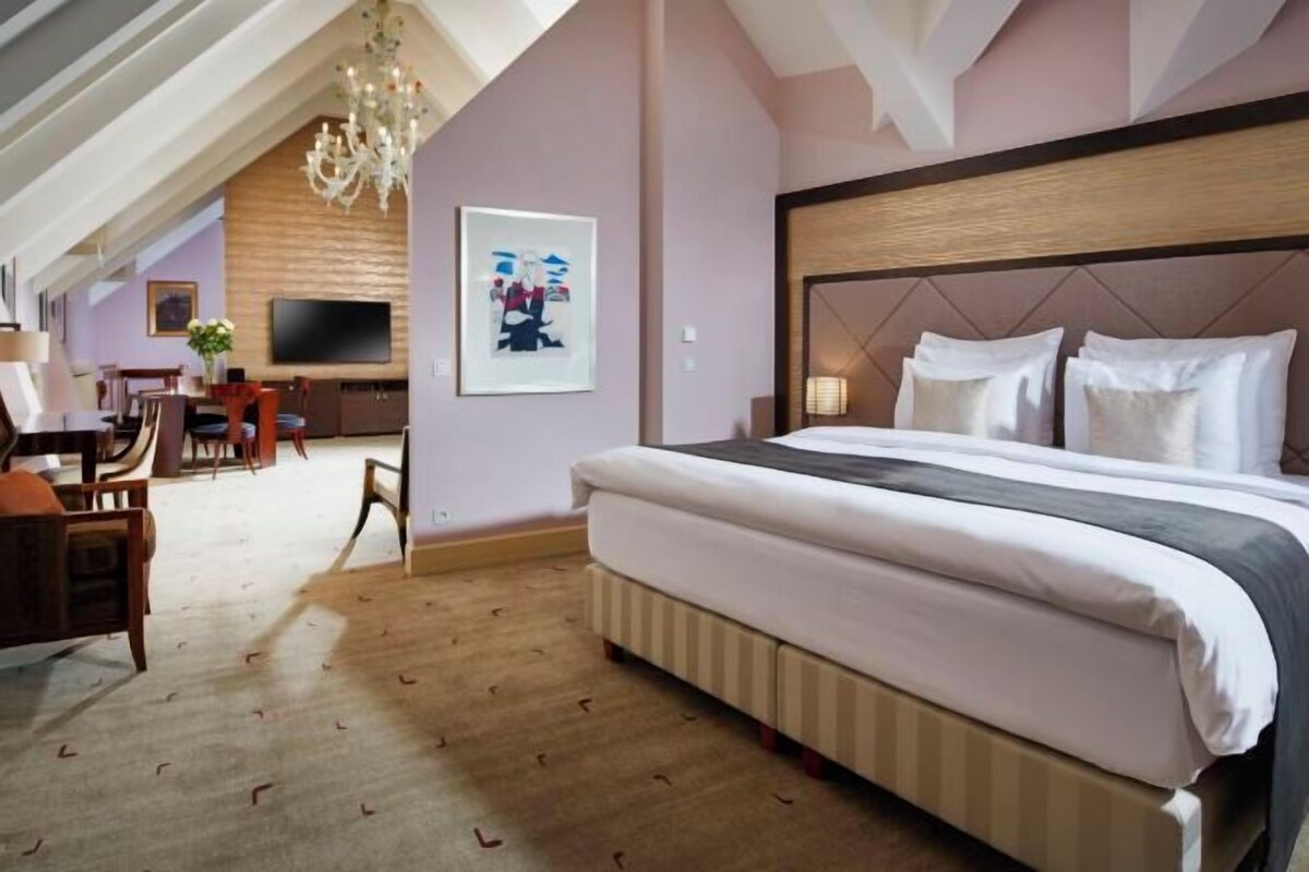A hotel room with a large bed, a television, a desk with a chair, and a chandelier.