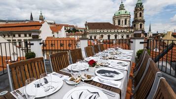 A rooftop dining table set with white plates, glasses, and cutlery, overlooking a cityscape with historic buildings and a church tower.