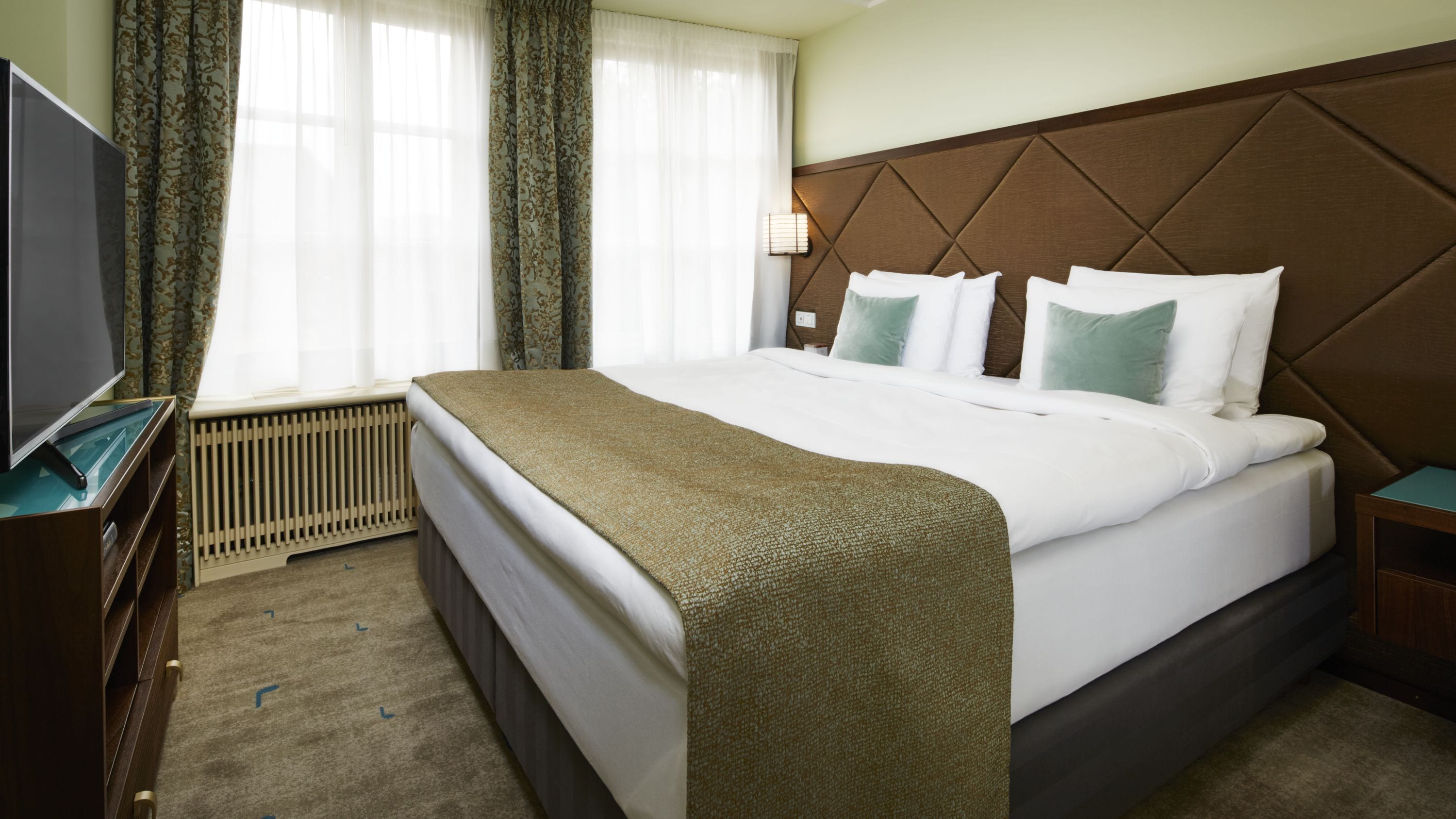 A hotel room with a bed, a television, a radiator, and curtains.