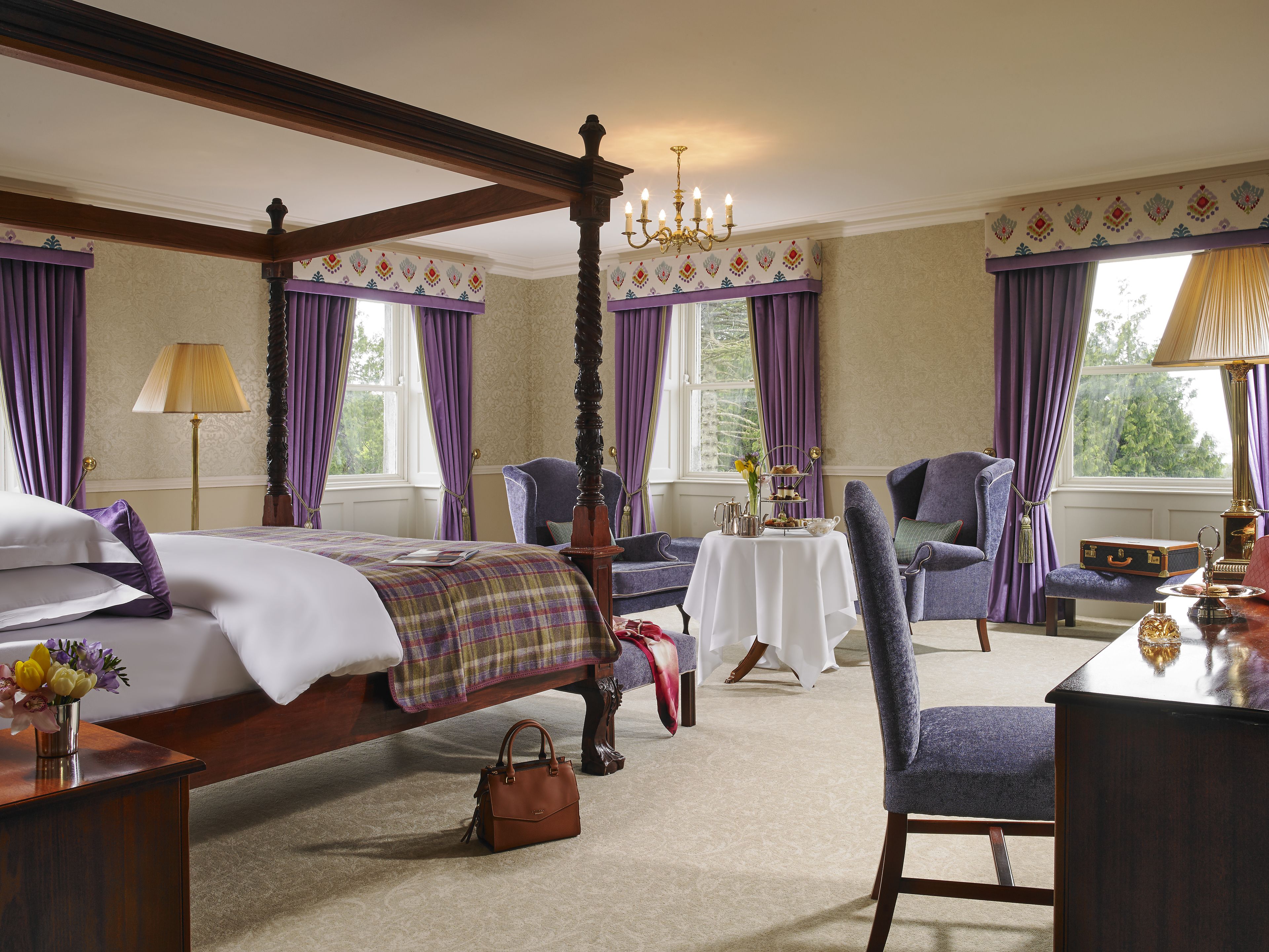 A four-poster bed with a plaid bedspread, a sitting area with a table and chairs, and a view of the outdoors through large windows.