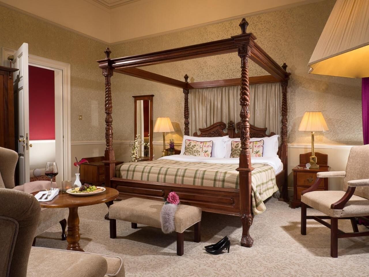 A four-poster bed with a plaid bedspread, two bedside tables with lamps, a bench with a fur throw, and a dining table with a plate of fruit and a glass of red wine.
