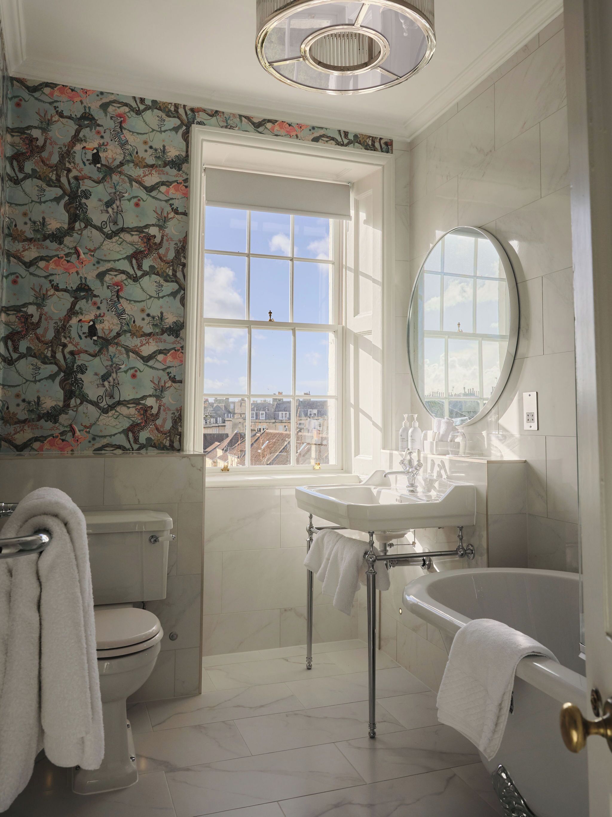 A bathroom with a toilet, sink, and bathtub, a window with a city view, and a decorative wallpaper with a nature theme.