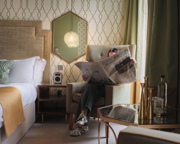 Une chambre d’hôtel avec un lit, une chaise et un journal, une table de chevet avec une radio et un miroir au mur.