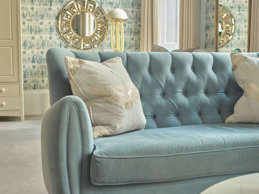 A light blue tufted sofa with white and gold pillows, a round gold mirror, and a gold floor lamp in a room with patterned wallpaper and a white door.