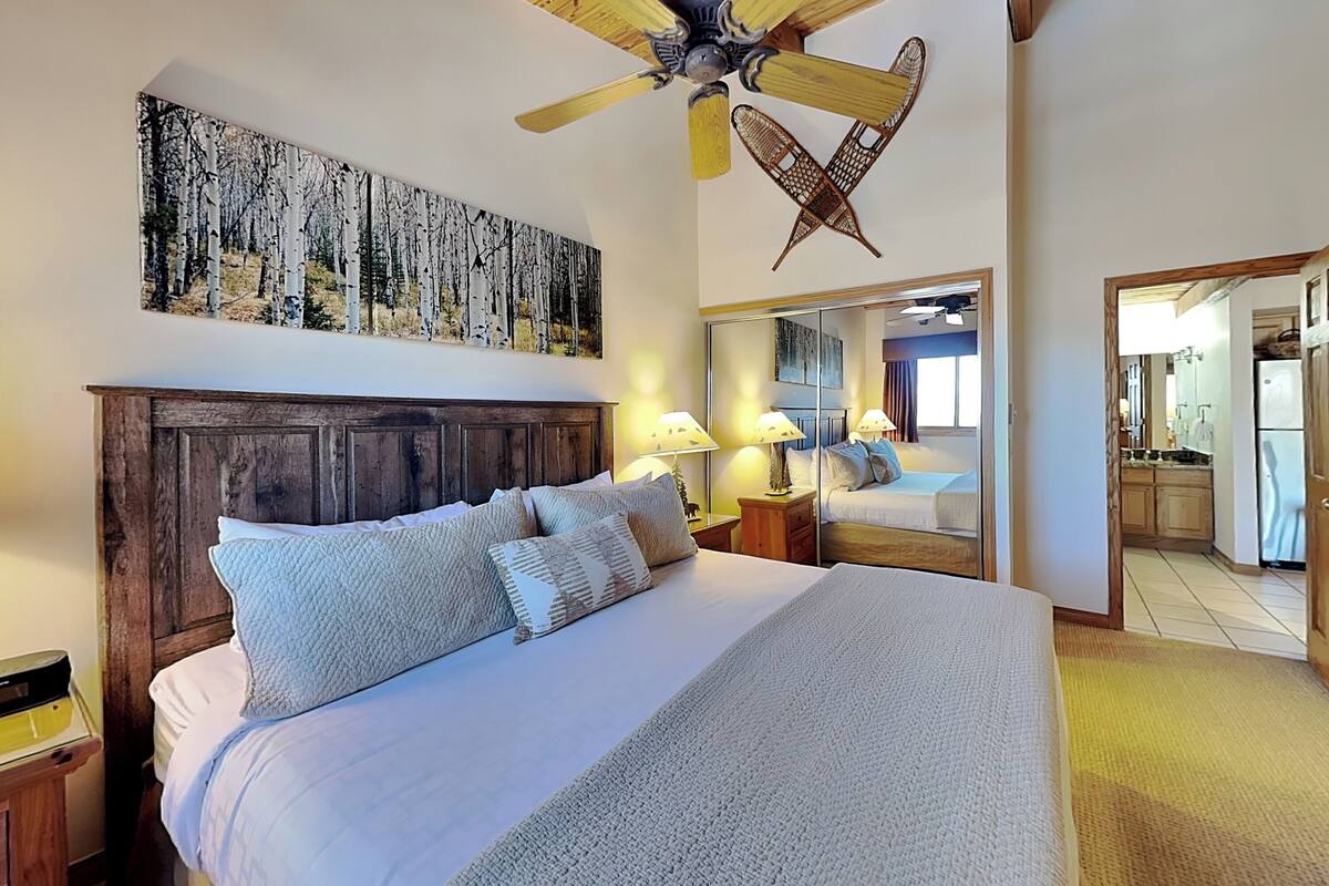 Une chambre à coucher avec une tête de lit en bois, un grand lit avec de nombreux oreillers, un ventilateur de plafond, un tableau accroché au mur et un miroir qui reflète la pièce.