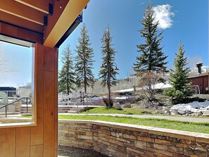 A view from a wooden-framed window overlooking a stone wall, a grassy area, and several tall evergreen trees. - The Timberline Condominiums (Snowmass Village)