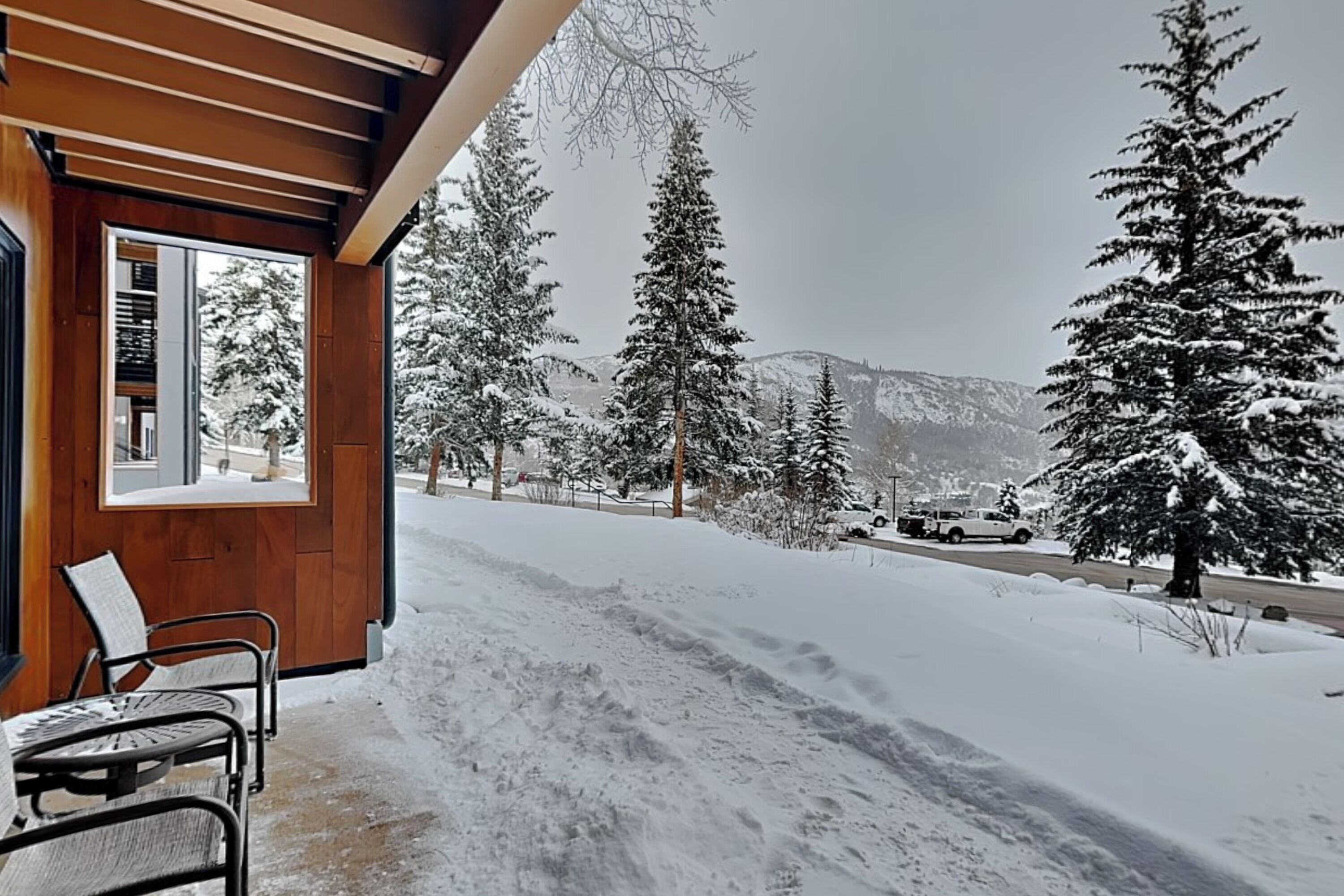 A wooden exterior with a covered porch, snow-covered ground, and a view of snow-laden trees and mountains.