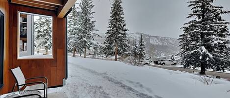 A wooden exterior with a covered porch, snow-covered ground, and a view of snow-laden trees and mountains.