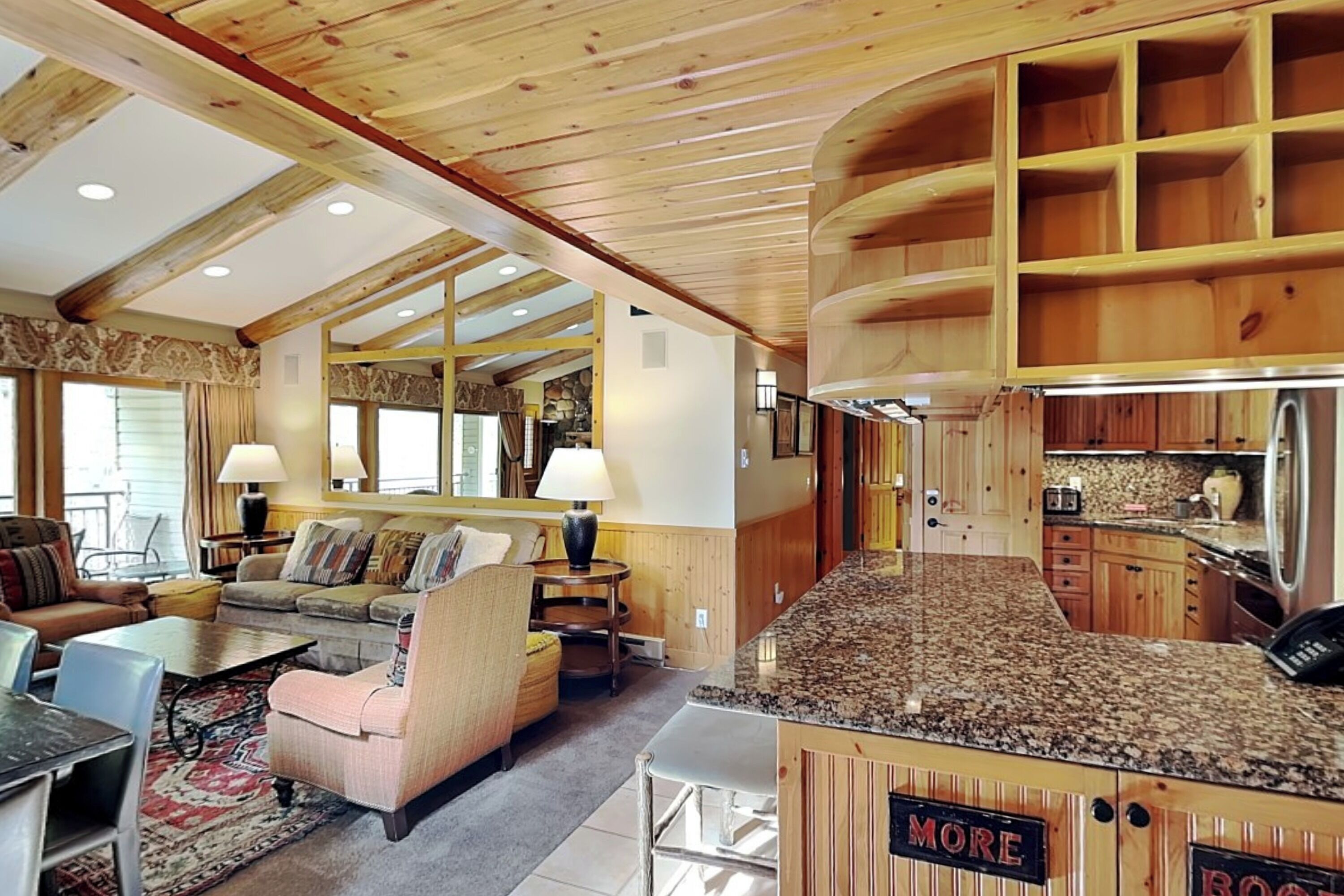 A spacious living room with a large sofa, a coffee table, and a dining area. There is a kitchen with wooden cabinets and a granite countertop.
