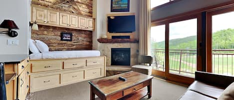 A cozy room with a bed, wooden cabinets, a fireplace, a coffee table, and a balcony with a view of the outdoors.