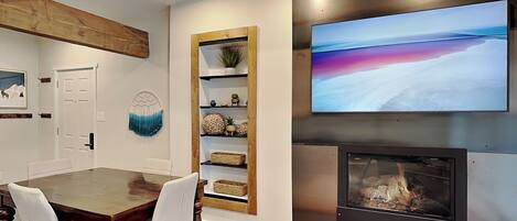 Un comedor con una mesa y sillas de madera, una chimenea, un televisor montado en la pared y un estante con objetos decorativos.