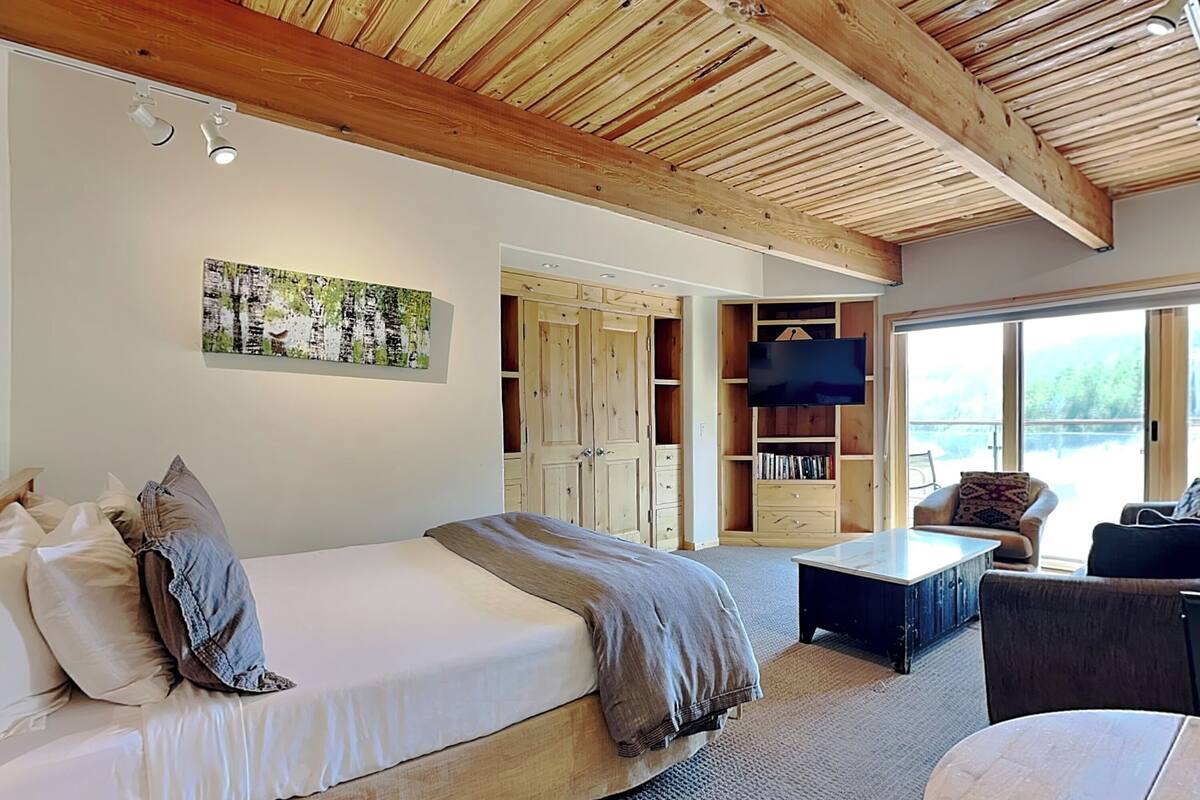 Une chambre avec un plafond en bois, un lit avec des oreillers, une télévision et une vue sur l’extérieur grâce à de grandes fenêtres.