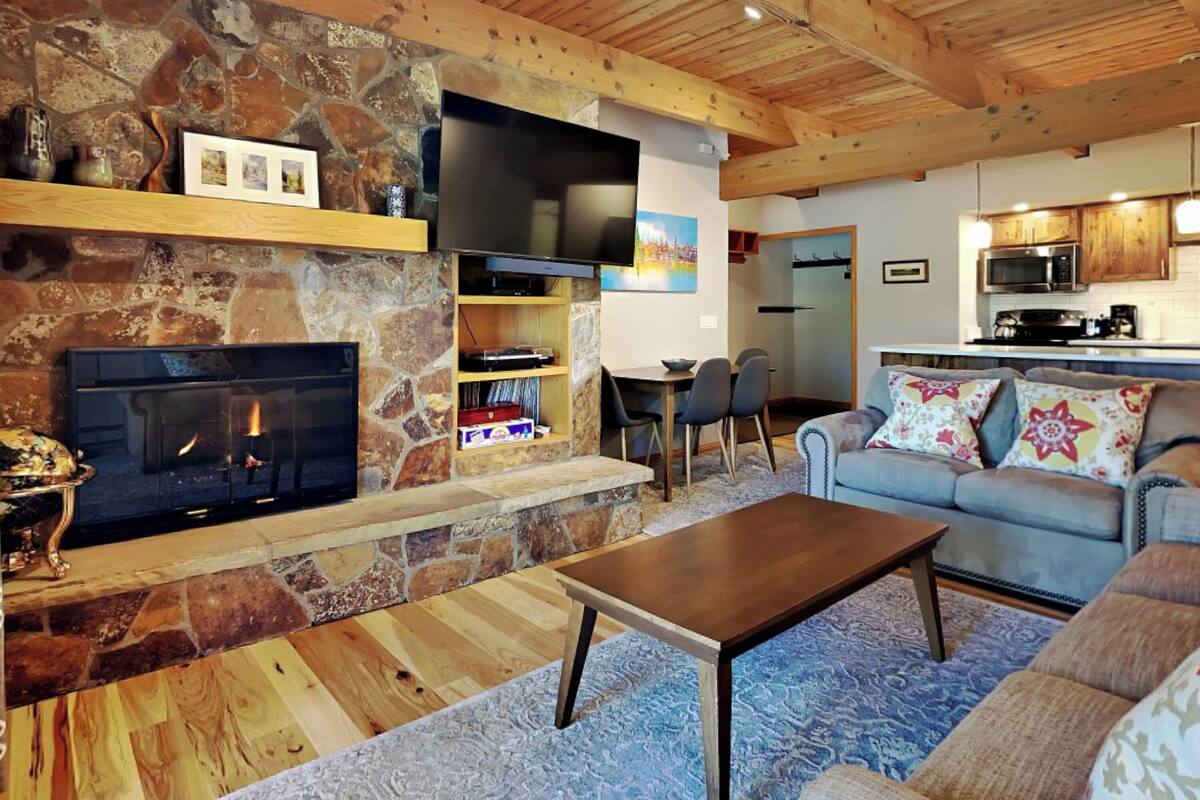 Un salon chaleureux avec une cheminée en pierre, un téléviseur à écran plat fixé au mur, un coin repas avec une table et des chaises, et une cuisine en arrière-plan.