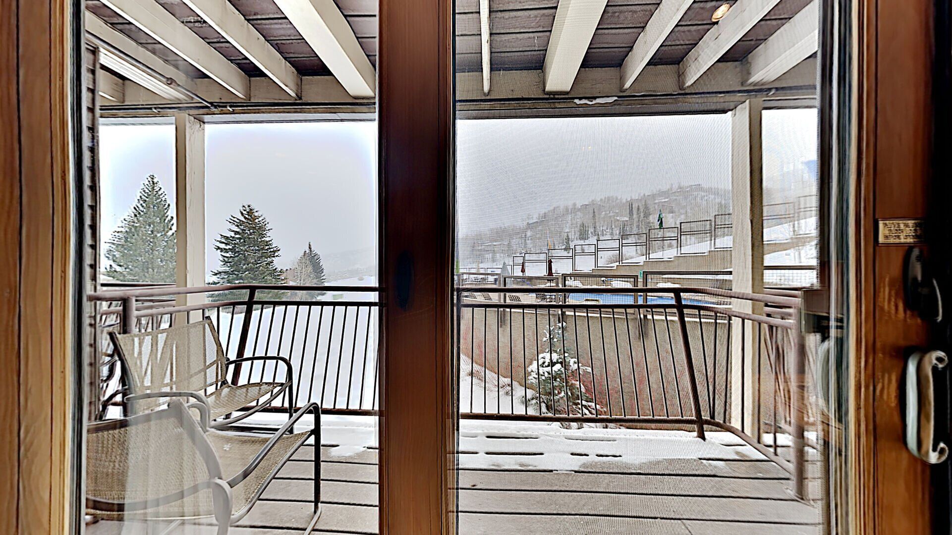 A balcony with a metal railing, a chair, and a view of snow-covered trees and a distant building.