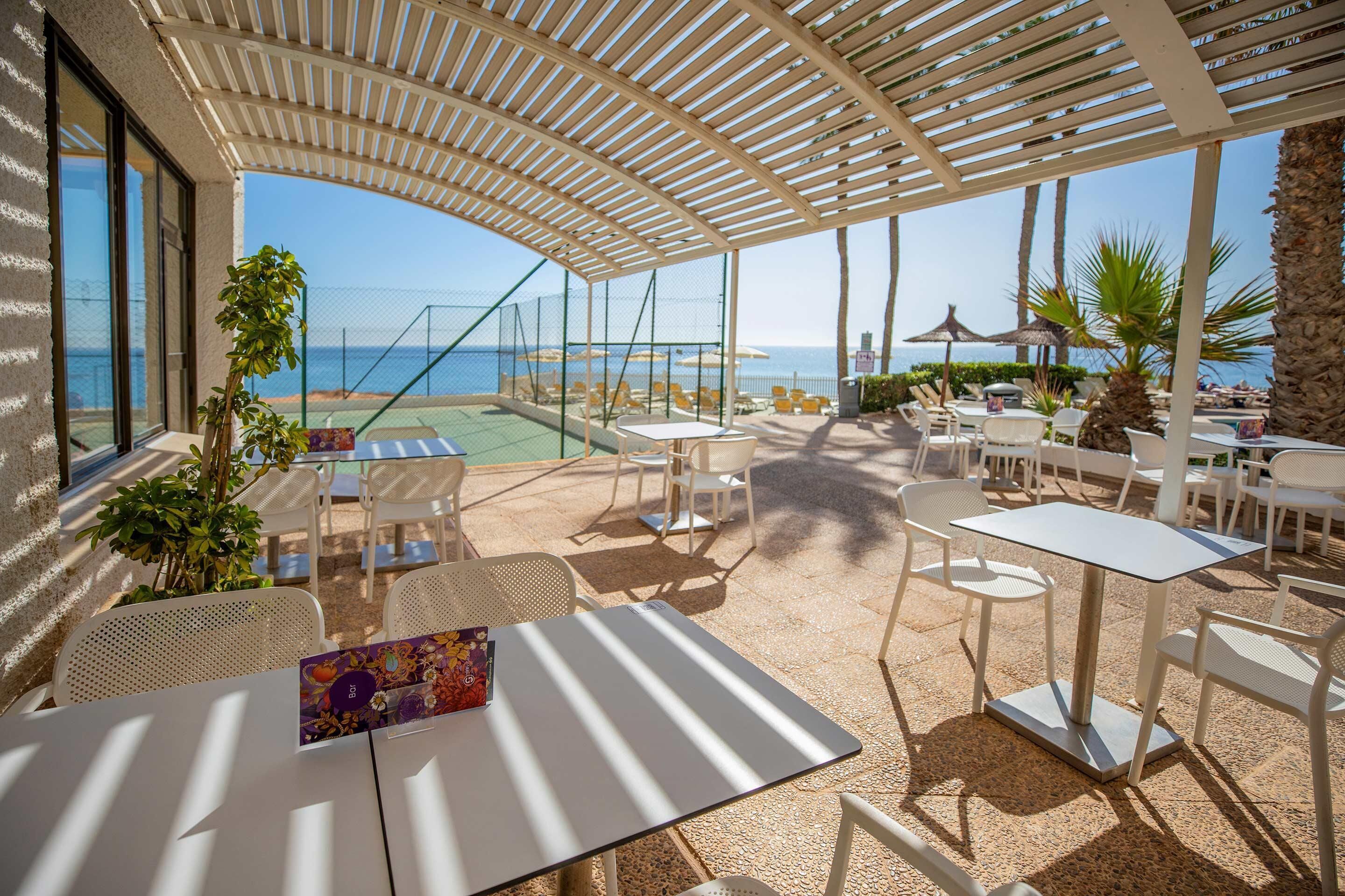 Un espacio de estar al aire libre techado con mesas y sillas, con vistas a una piscina y una playa.