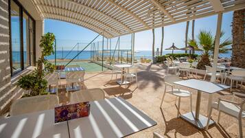 A covered outdoor seating area with tables and chairs, overlooking a pool and beach.