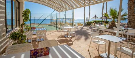 Un espacio de estar al aire libre techado con mesas y sillas, con vistas a una piscina y una playa.