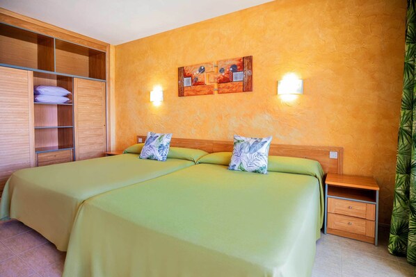 Una cama doble con ropa de cama verde y almohadas estampadas, un armario de madera, una mesita de noche con una lámpara y una obra de arte en la pared.