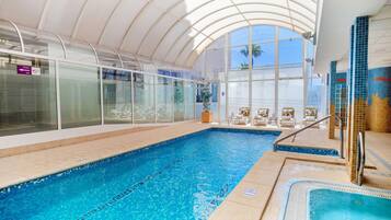 Una piscina interior con paredes de vidrio transparente, un ventanal grande y una zona de solárium con baldosas.