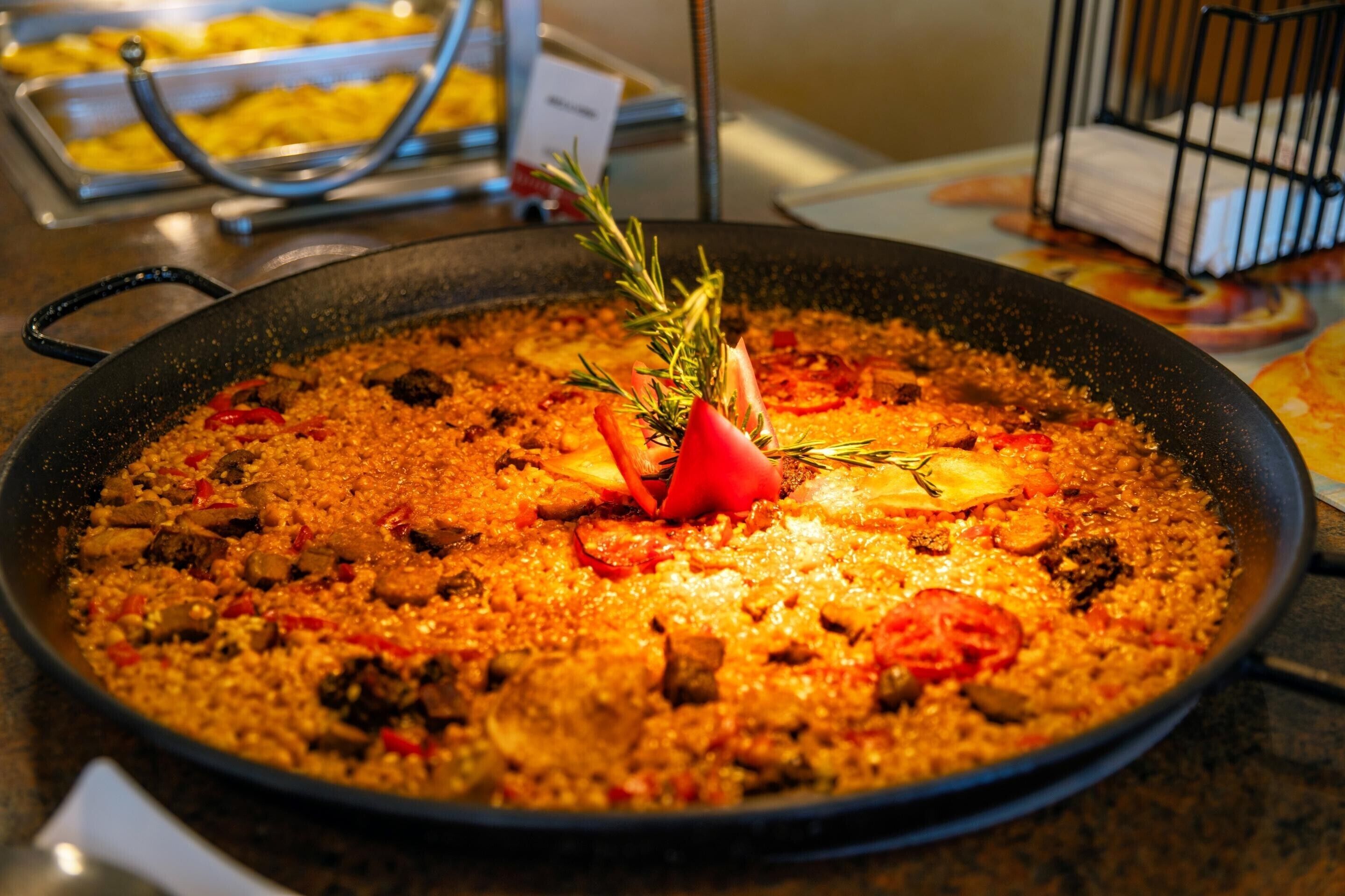 Una sartén grande, redonda y negra llena de un plato con arroz, carne y verduras, adornado con una ramita de romero.