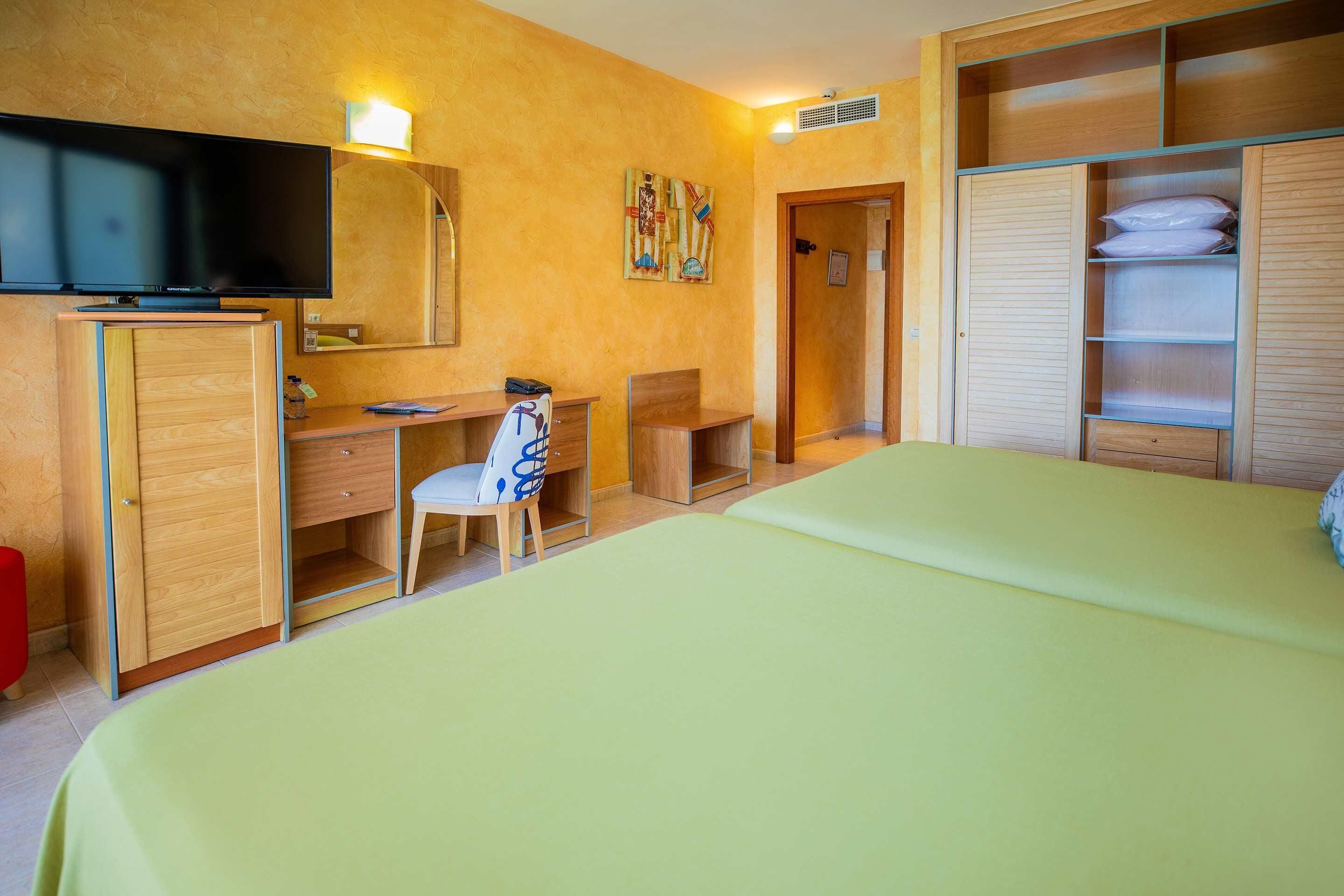 Habitación de hotel con cama, escritorio, silla, televisor, armario y espejo.