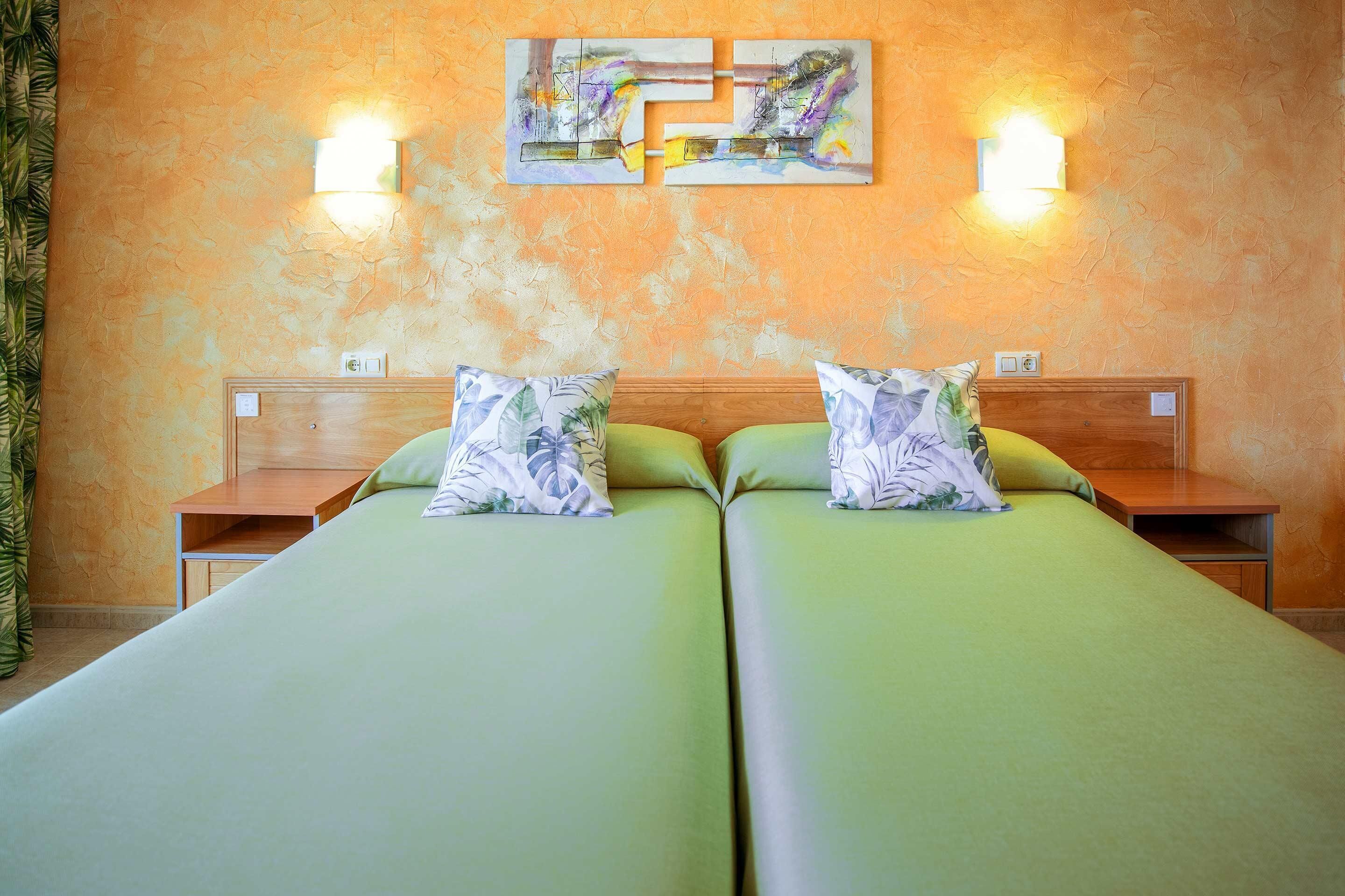 Ein Zimmer mit zwei Betten, jeweils mit Kissen und Nachttisch. An den Wänden sind Leuchten angebracht, und über den Betten hängt ein Gemälde.