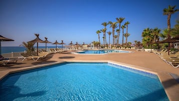 A pool area with multiple sun loungers and palm trees, surrounded by a fence, with a clear view of the sea in the background.