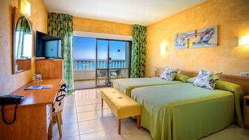 Habitación de hotel con balcón, un escritorio con silla, cama con ropa de cama verde y un televisor sobre un soporte de madera.