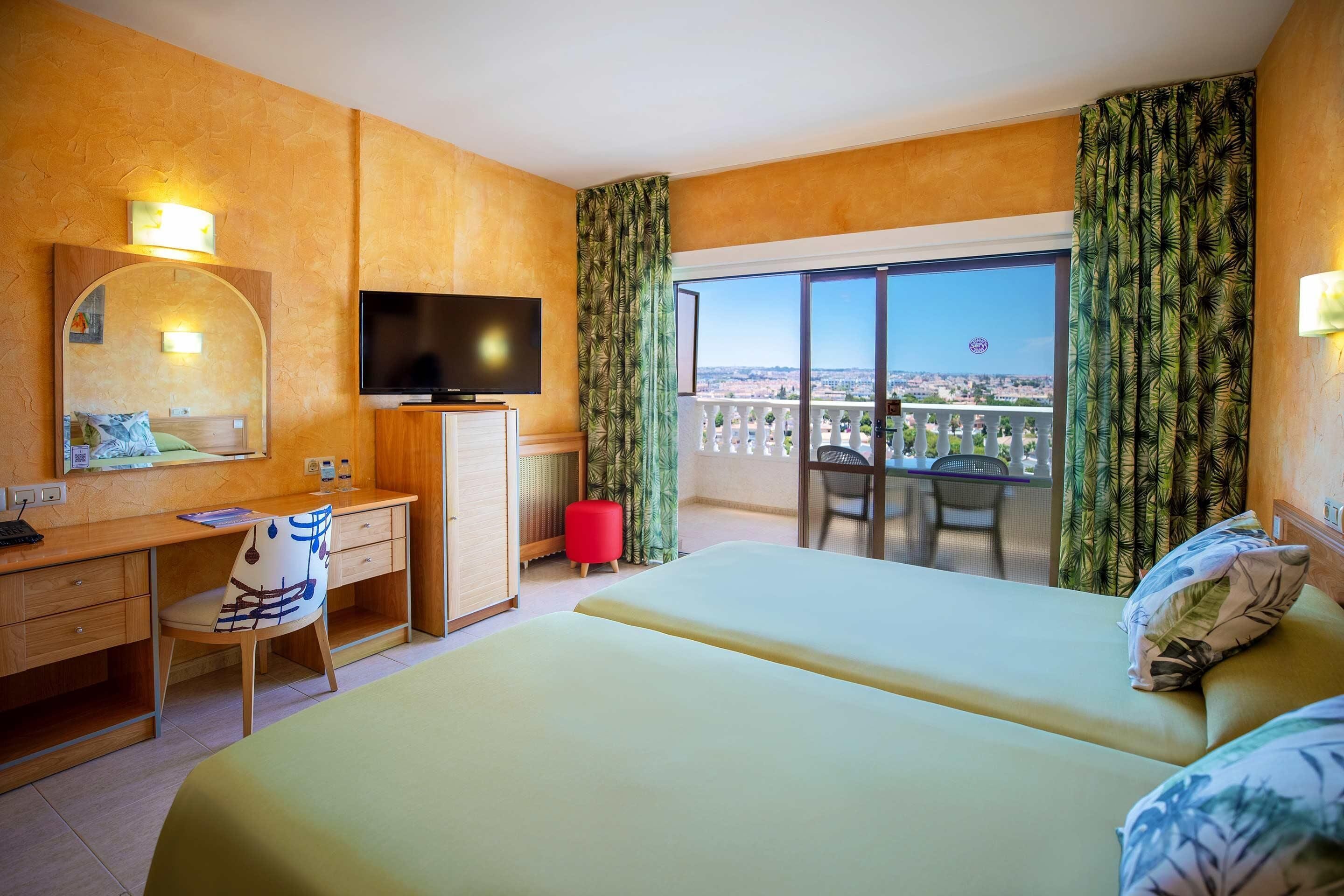 A hotel room with a bed, desk, chair, TV, and balcony with curtains.