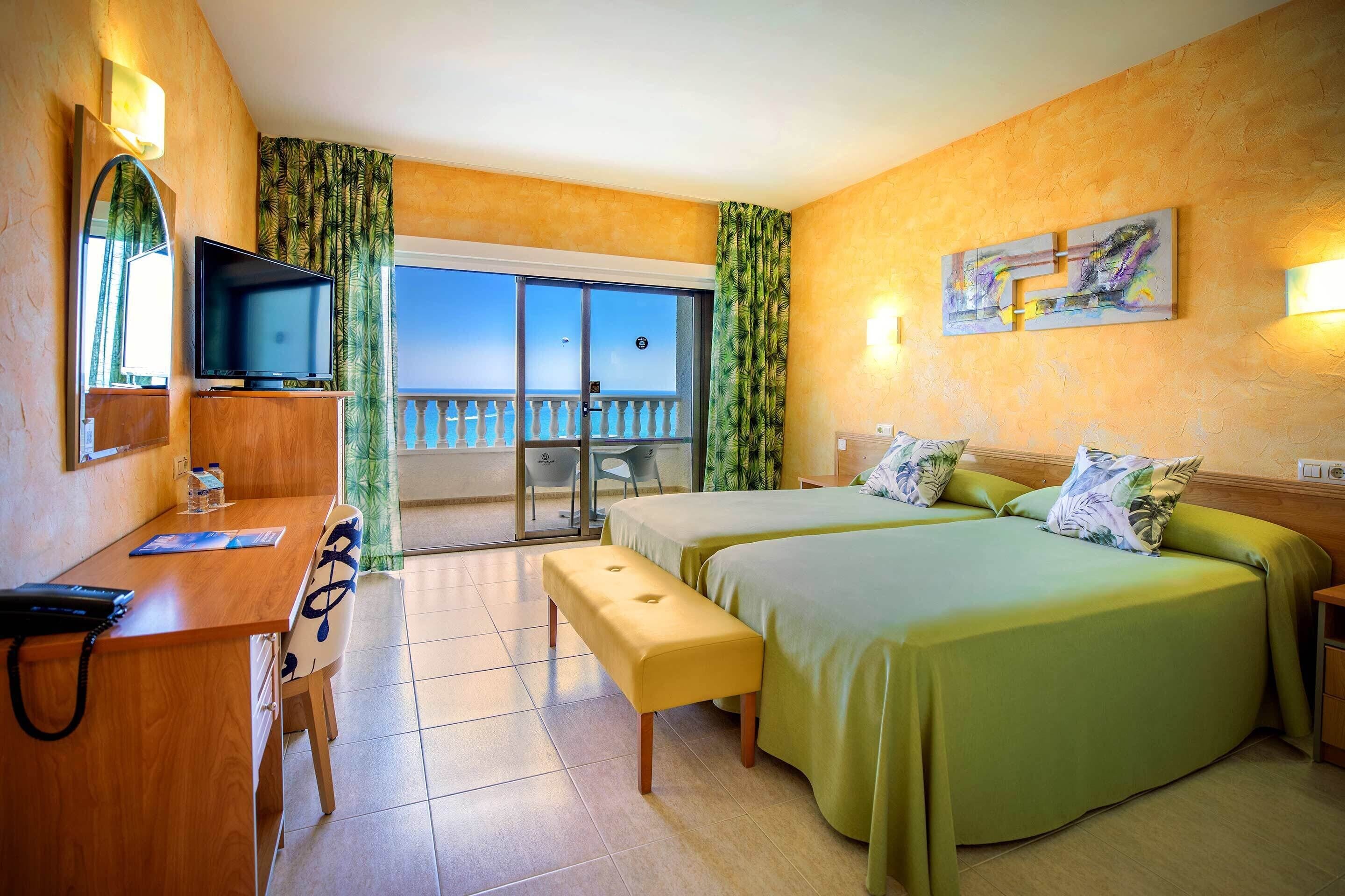 A hotel room with a balcony, a desk with a chair, a bed with green bedding, and a television on a wooden stand.