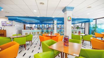 Un restaurante muy iluminado con asientos verdes y anaranjados, una zona de bar al fondo y un diseño de techo moderno.
