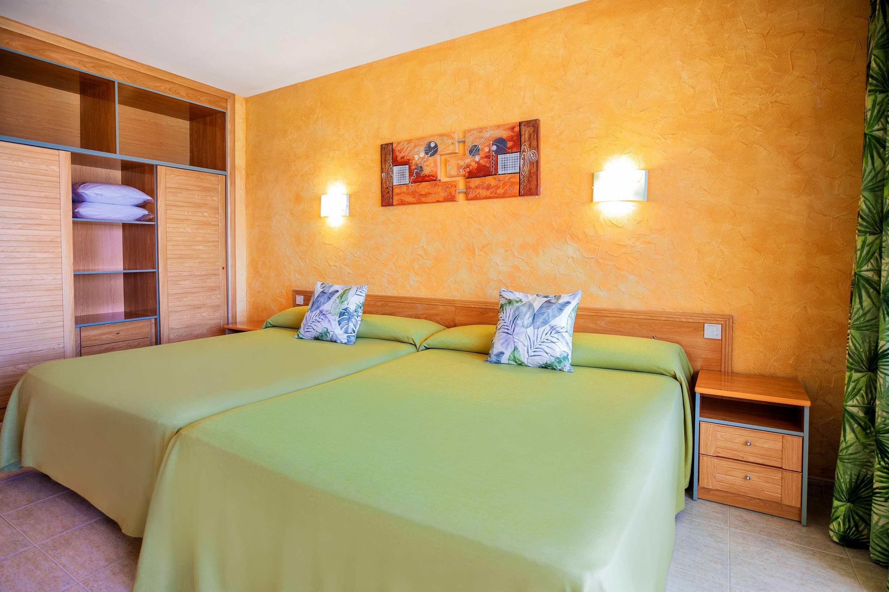 A double bed with green bedding, a wooden wardrobe, a nightstand, and a wall-mounted light fixture.