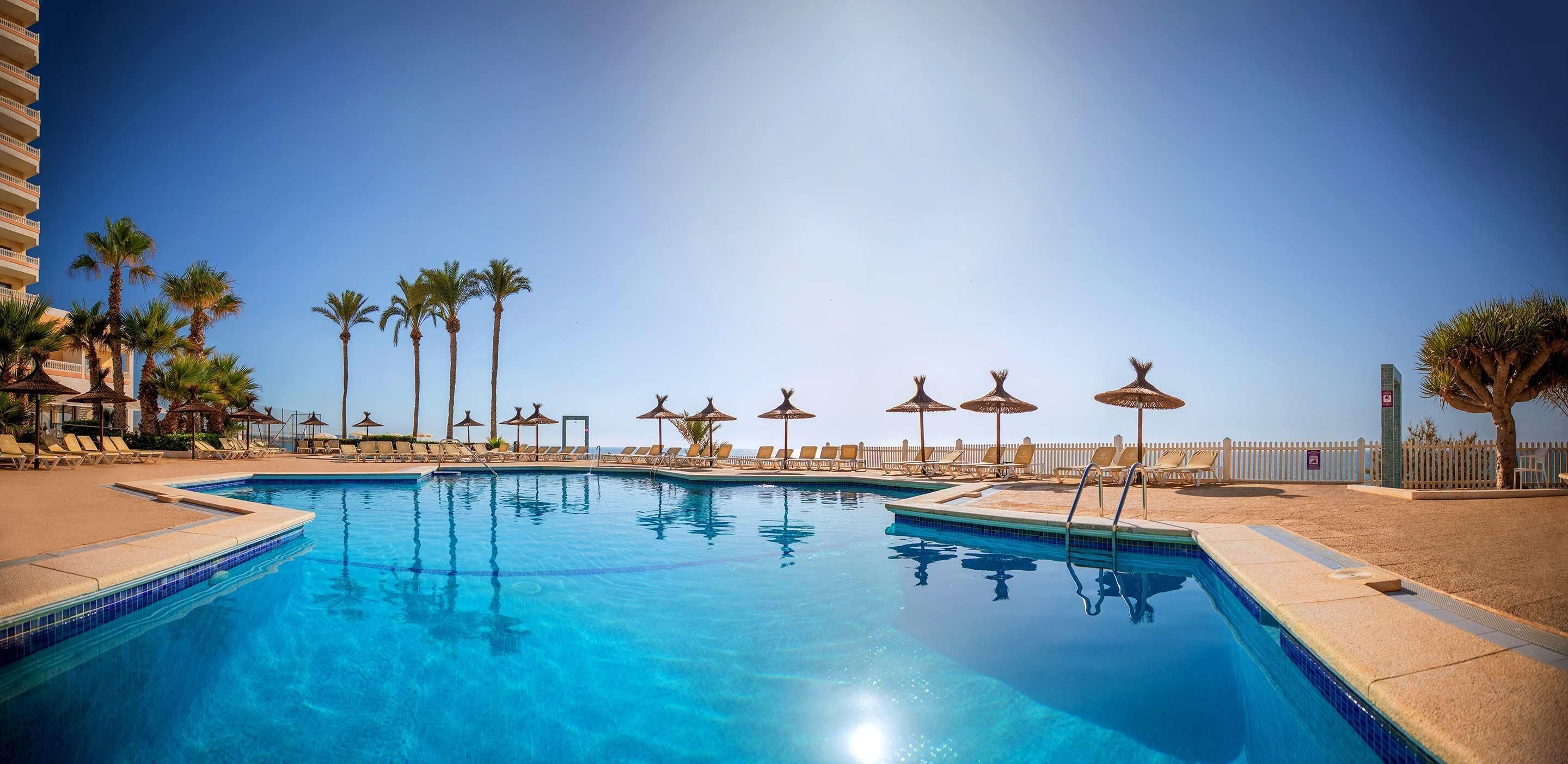 A clear blue pool with lounge chairs and palm trees, surrounded by a white fence.