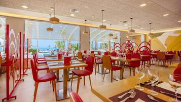 Un restaurante con varias mesas y sillas, amplias ventanas que dan a la piscina y palmeras, e iluminación colgante.