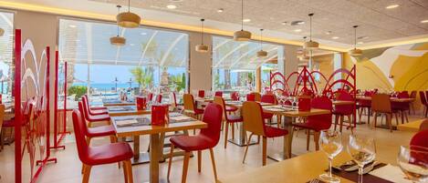 A restaurant with multiple dining tables and chairs, large windows offering a view of the pool and palm trees, and pendant lighting.