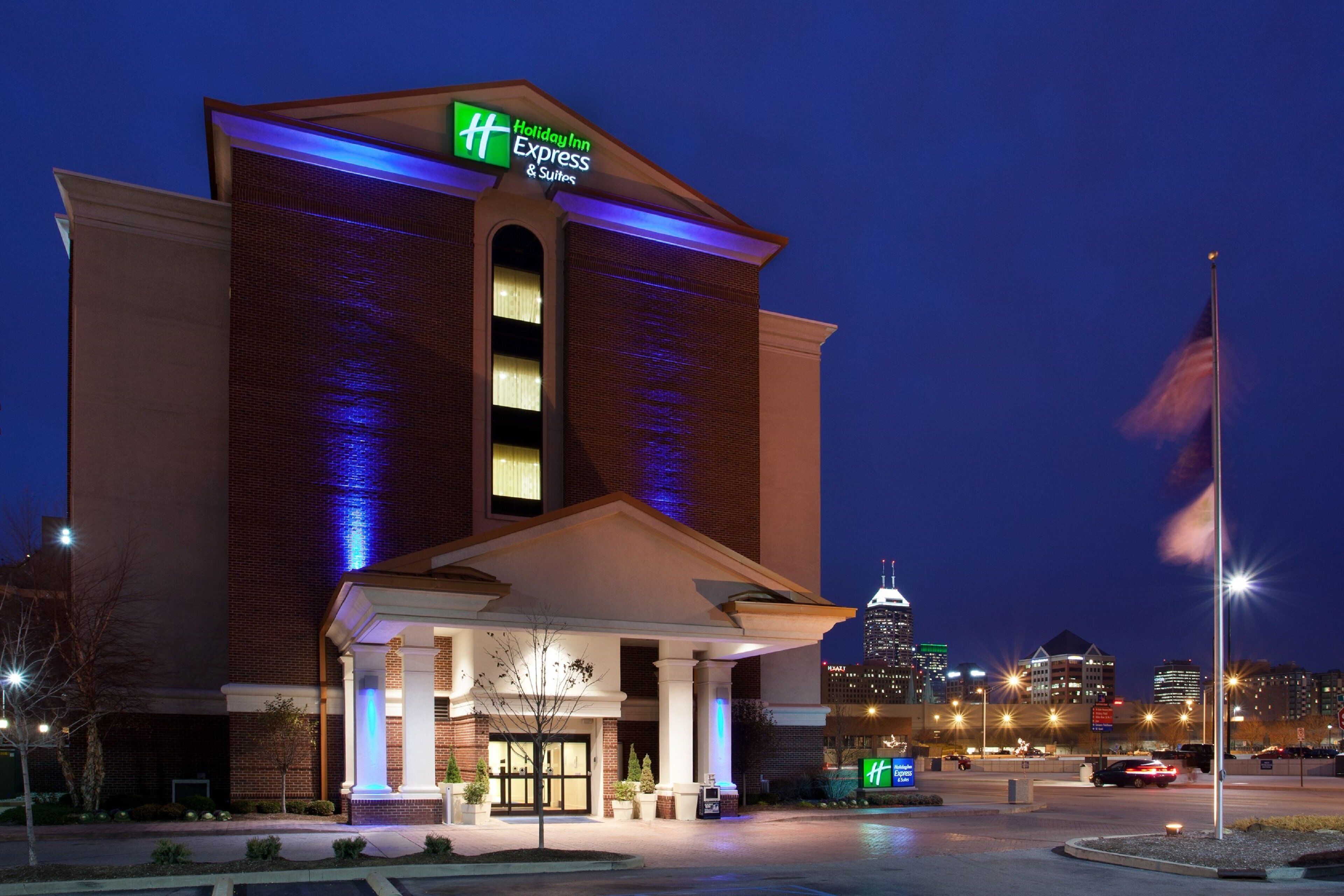 A Holiday Inn Express & Suites hotel with a prominent sign, a well-lit entrance, and a view of the city skyline in the background.