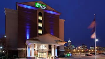 A Holiday Inn Express & Suites hotel with a prominent sign, a well-lit entrance, and a view of the city skyline in the background.