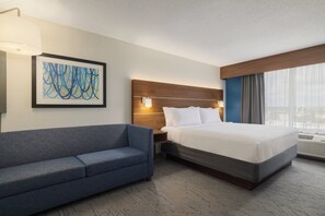Une chambre d’hôtel comprenant un lit, un canapé, une table de chevet, une lampe fixée au mur et un tableau abstrait encadré.