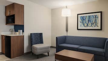 A modern hotel room with a kitchenette, a grey sofa, a blue armchair, a wooden coffee table, and a framed abstract painting on the wall.